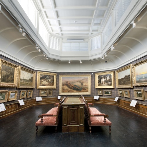 Una galería de arte con grandes pinturas enmarcadas en las paredes, una vitrina central y bancos tapizados en rojo.