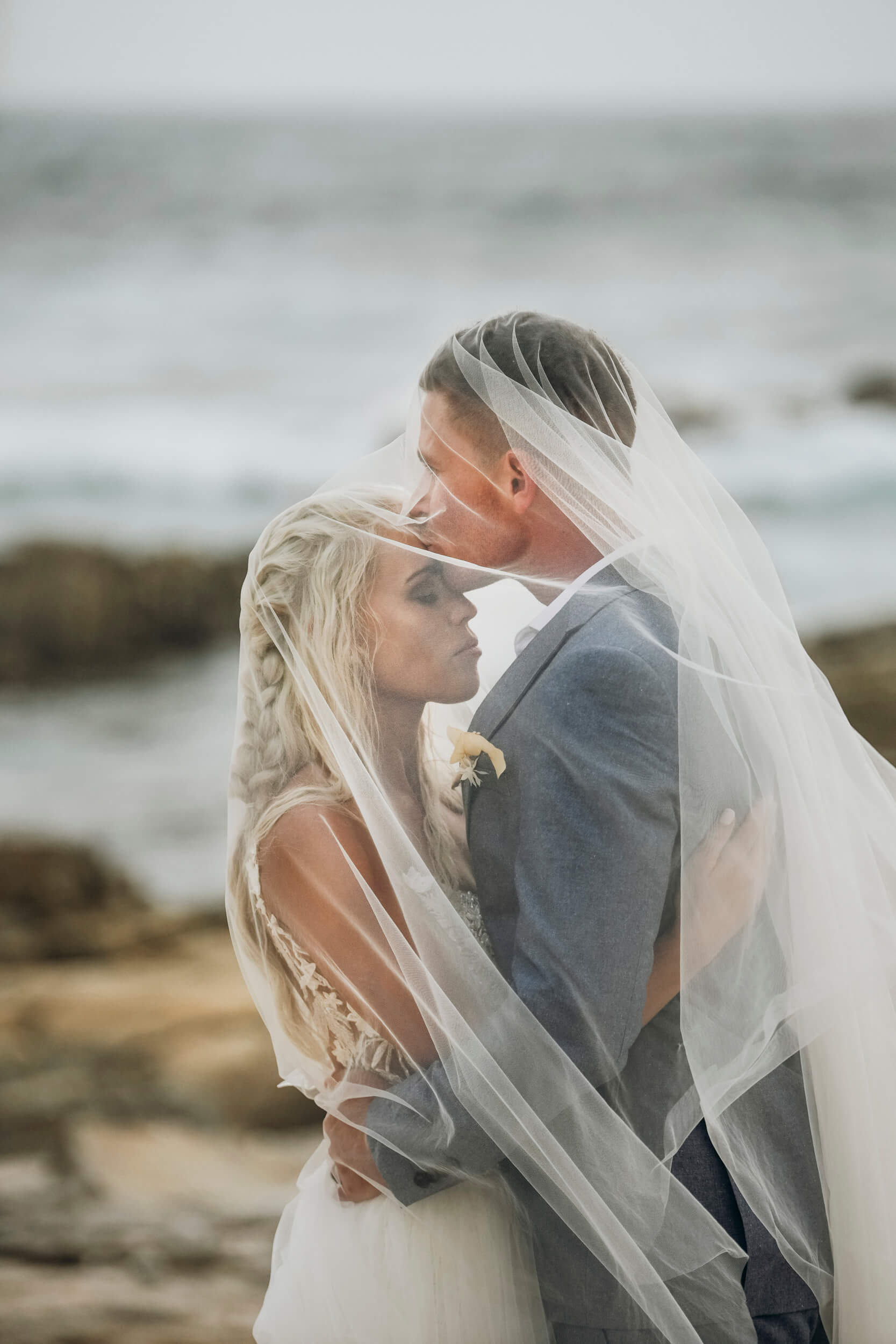 Bride and groom under windswept veil during coastal wedding portrait session