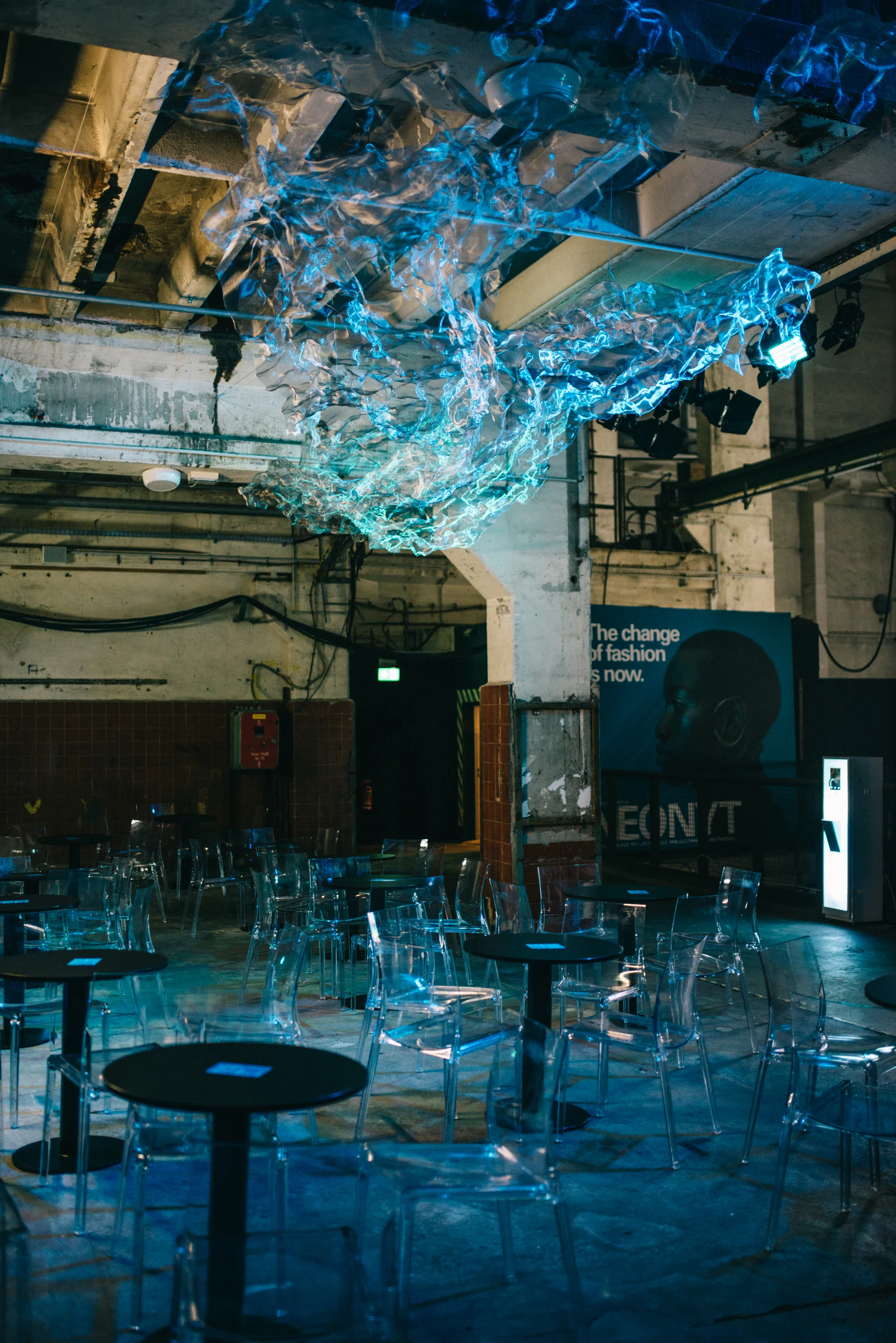 Floating sculpture of translucent foil glowing blue above black bistro tables and transparent chairs at FASHIONTECH Berlin.