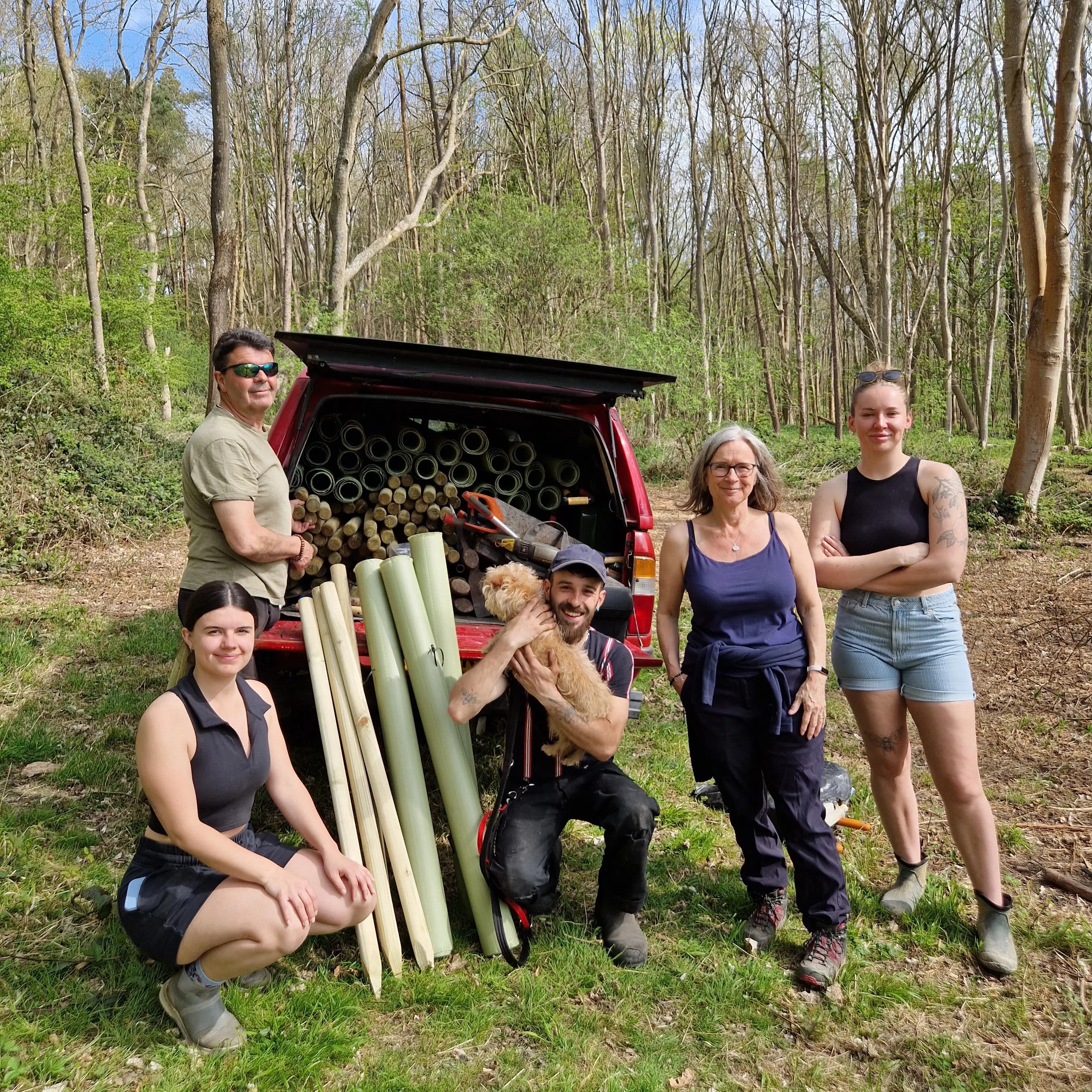 A group of six people stands in a wooded area, with some holding wooden planks and a truck in the background.