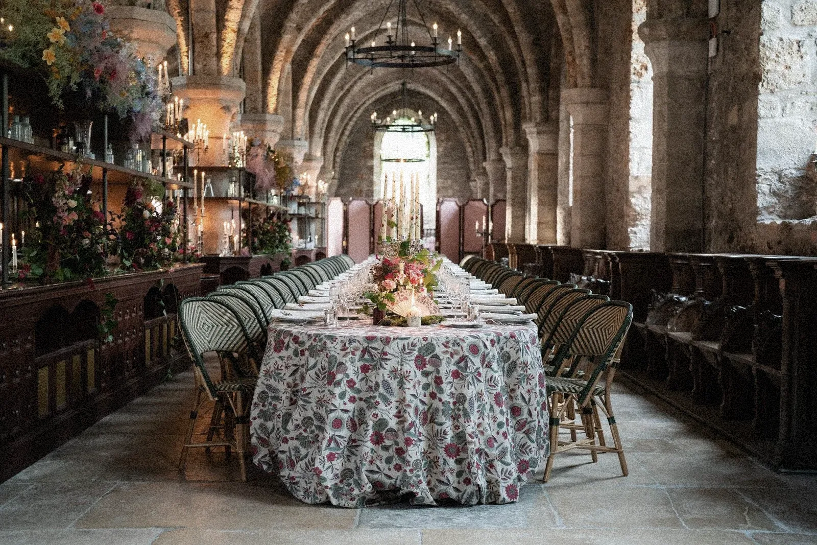 Long banquet table adorned with floral tablecloth and candles inside a historic stone hall with vaulted ceilings and warm, romantic lighting.