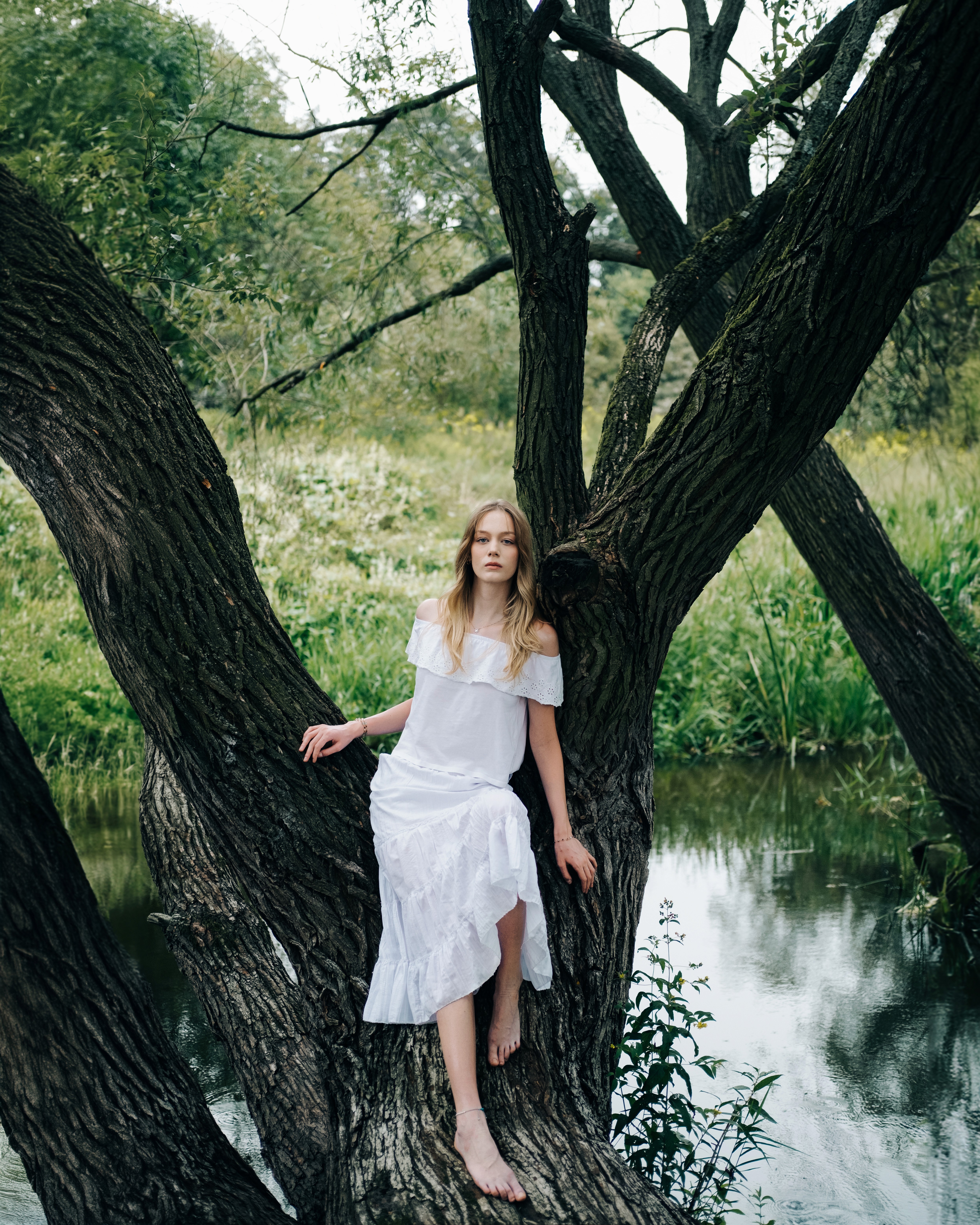 Hania – Cold Eden – seated barefoot in the fork of a tree near a quiet river, surrounded by greenery