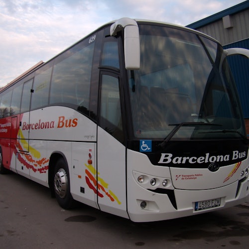 A white and red bus labeled "Barcelona Bus" parked on a paved surface near a blue industrial building.