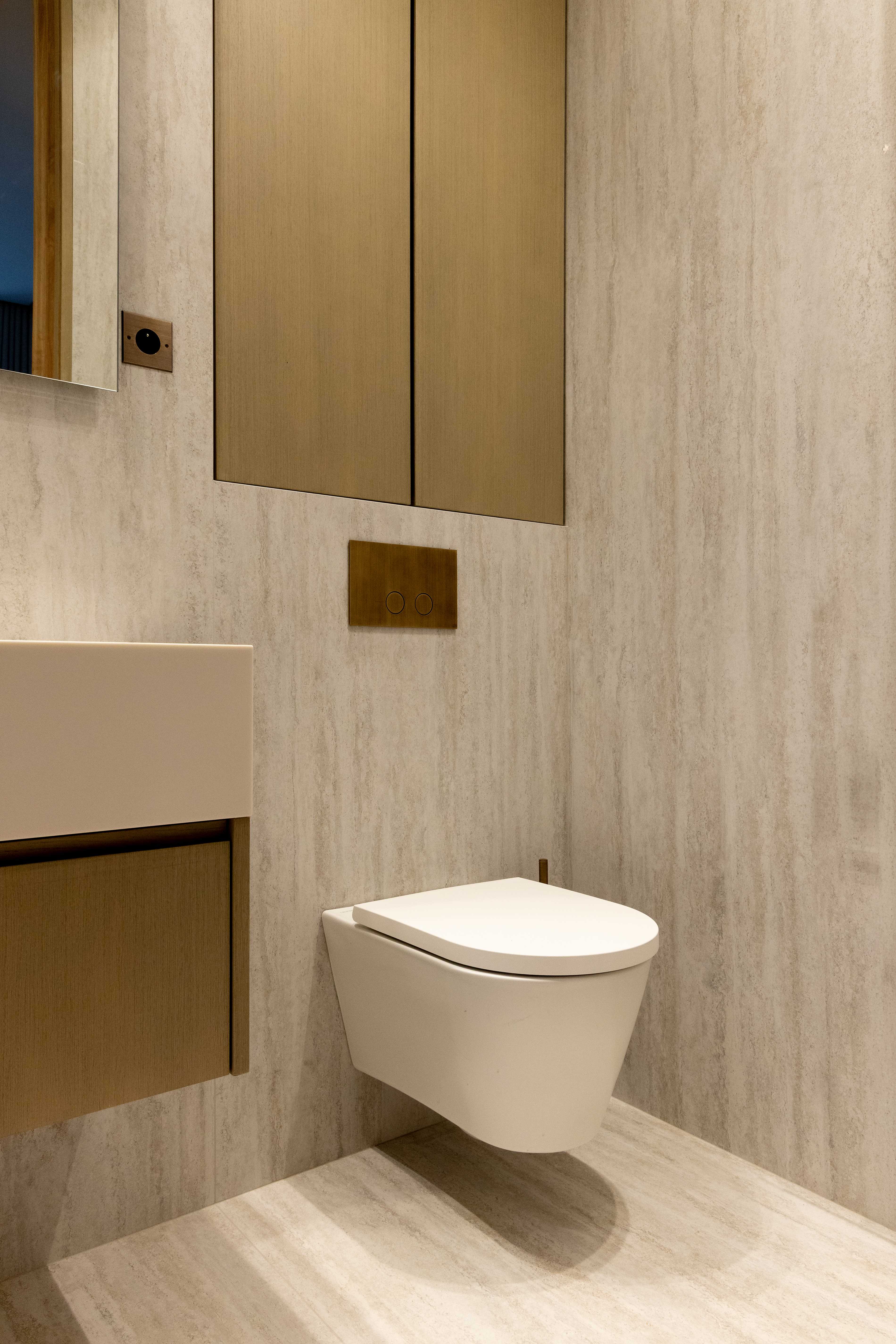 Toilettes suspendues blanches avec plaque de chasse doree, placards bois clair et murs travertin