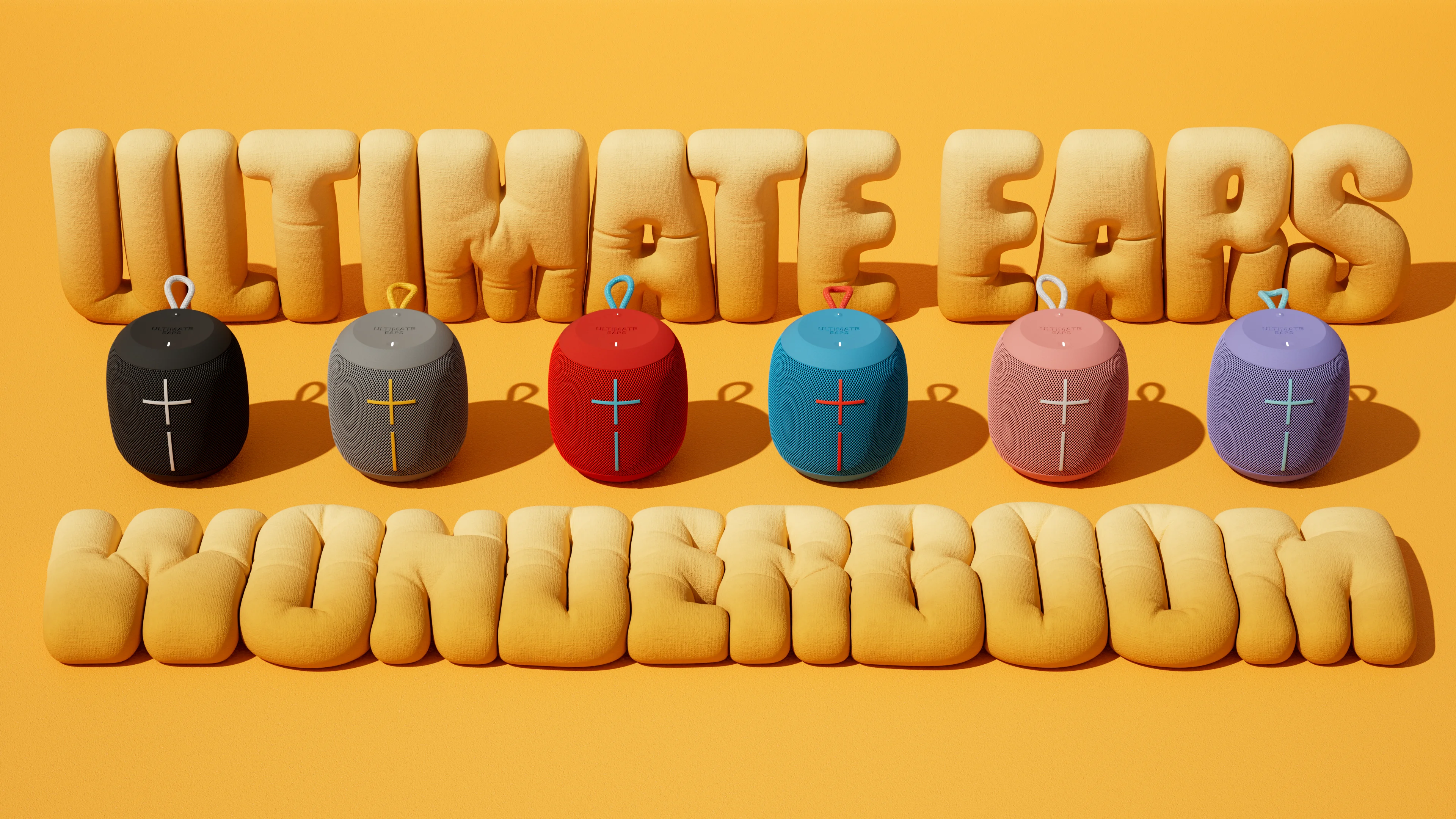 Six Ultimate Ears Wonderboom speakers in black, grey, red, blue, pink and purple lined up in front of inflated yellow Ultimate Ears Wonderboom lettering