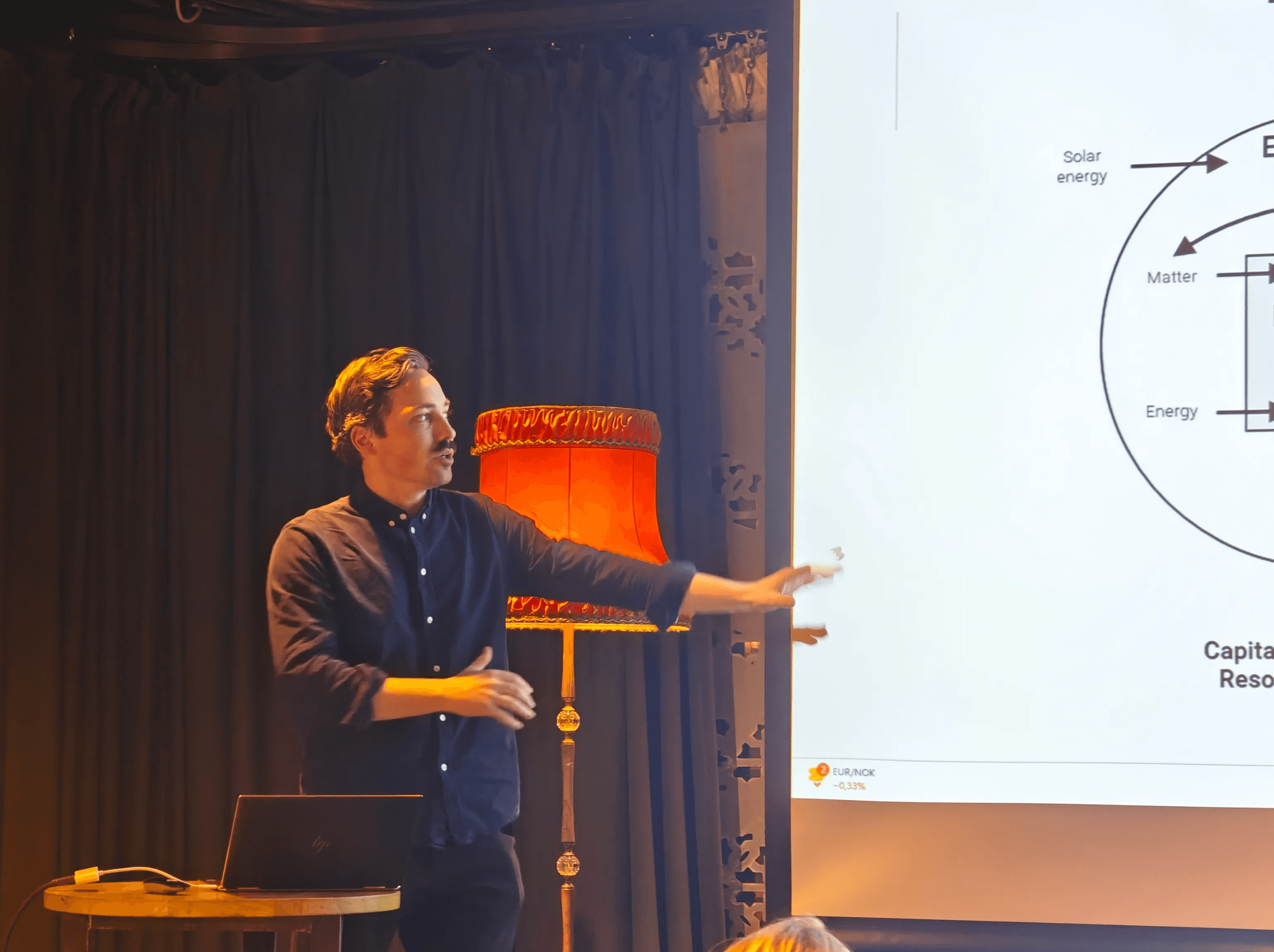 A person speaking on a stage showing something that is prejected onto a screen