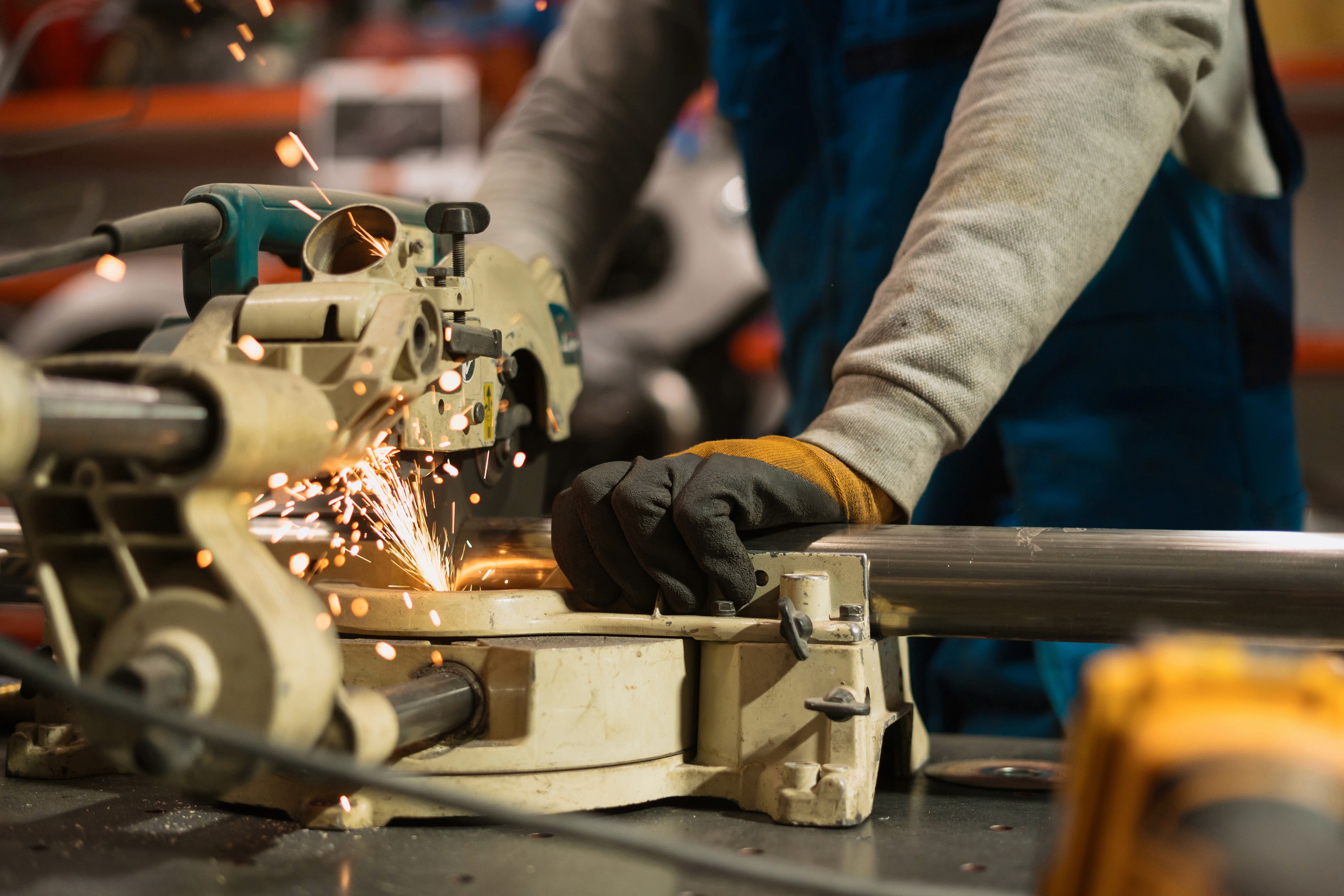 A person in work gloves uses a miter saw to cut a metal pipe, causing bright sparks. The setting is an industrial workshop, conveying focus and precision.
