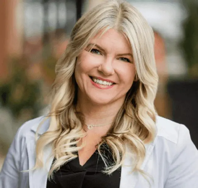 Smiling person with long, wavy blonde hair, wearing a black top and light jacket, set against a blurred background.
