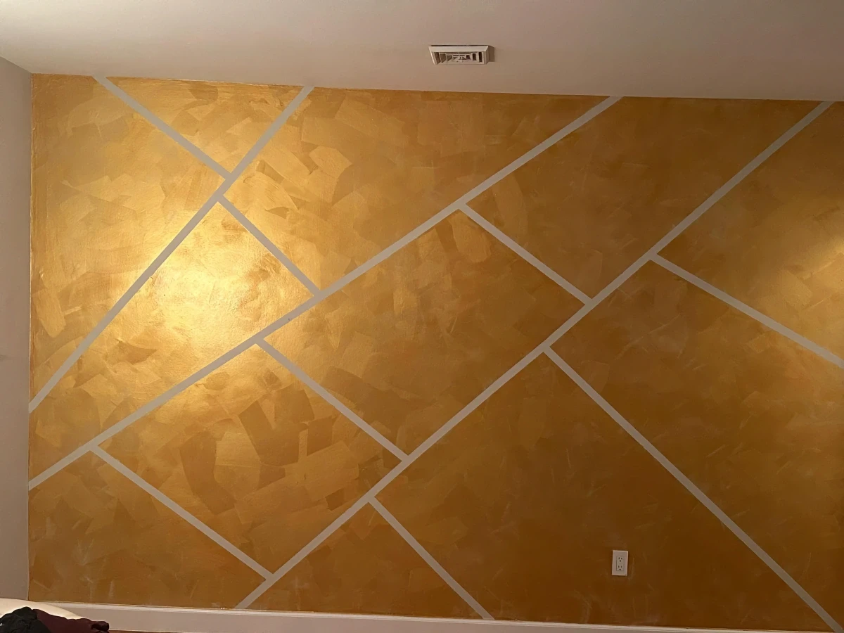 Accent wall finished in golden Venetian plaster with intersecting light gray diagonal stripes, creating a bold geometric design with a rich, textured sheen.