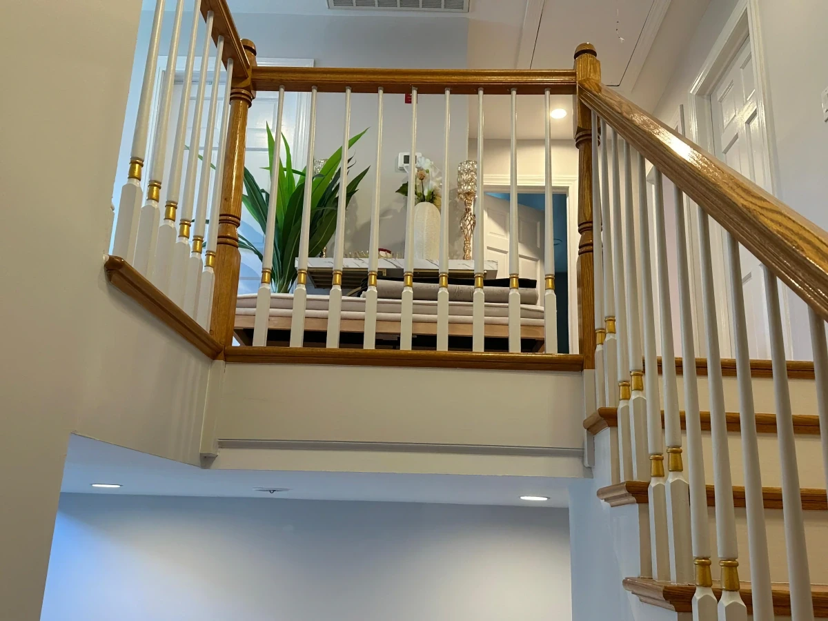 Staircase landing with white balusters, gold accents, and natural wood handrails, showcasing clean craftsmanship and a decorative upstairs hallway with plants and soft lighting.