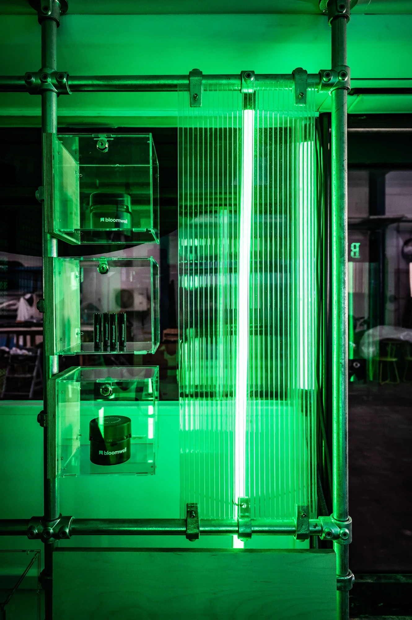 Detail of the Bloomwell bus interior with a glowing green-lit acrylic display case, vertical light bar and reflective product compartments inside scaffold frames.