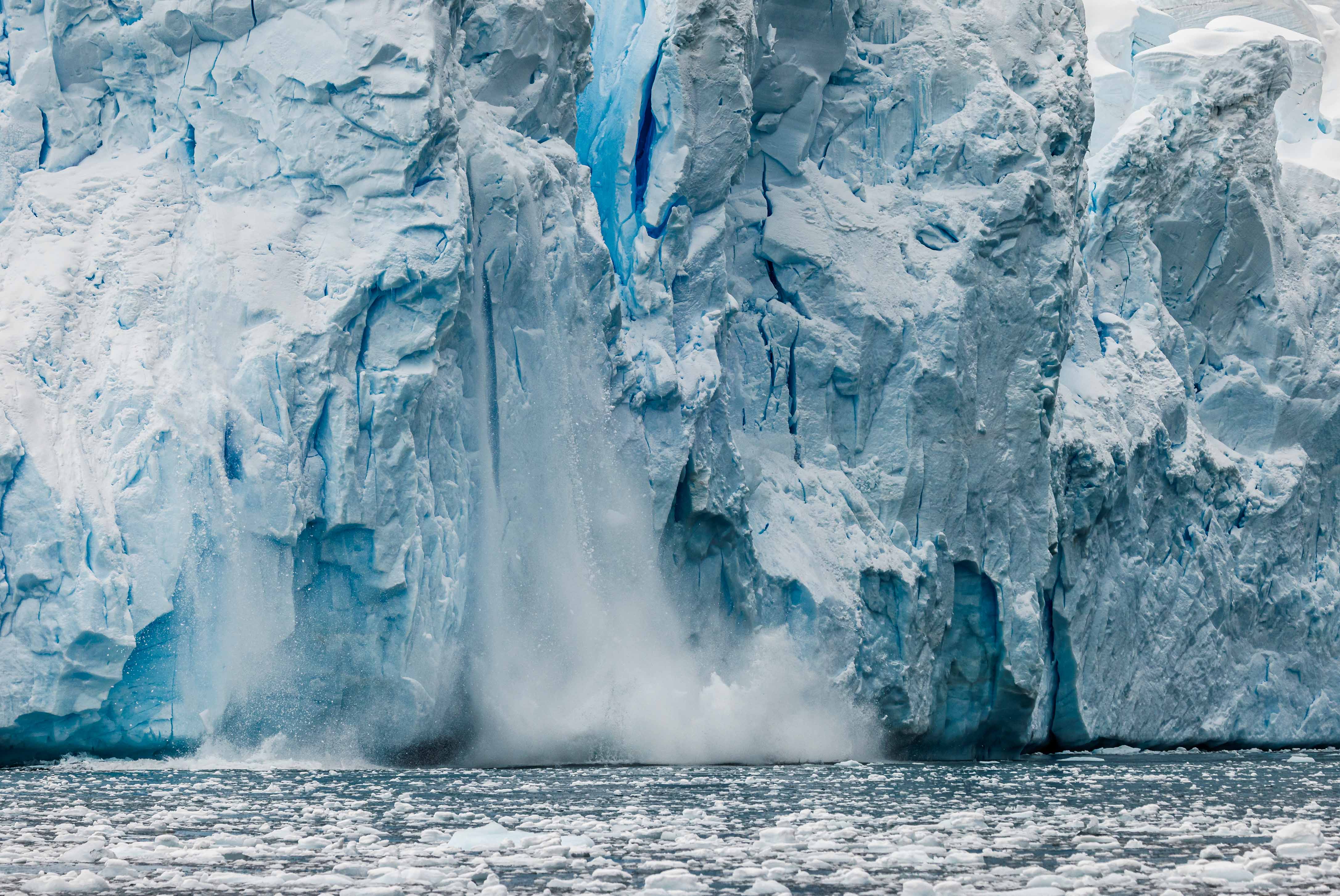 The photograph captures a dramatic scene of a glacier calving, where a large chunk of ice is breaking off and crashing into the water below. The glacier is massive, with towering, jagged ice formations that dominate the frame.