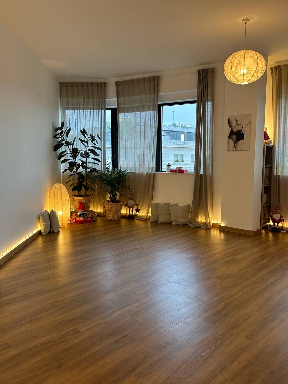 A wide shot of the empty studio space in the evening, illuminated by a warm paper pendant light and floor-level LED strips. The room features large potted plants, sheer beige curtains covering large windows, and subtle holiday decorations on the floor.