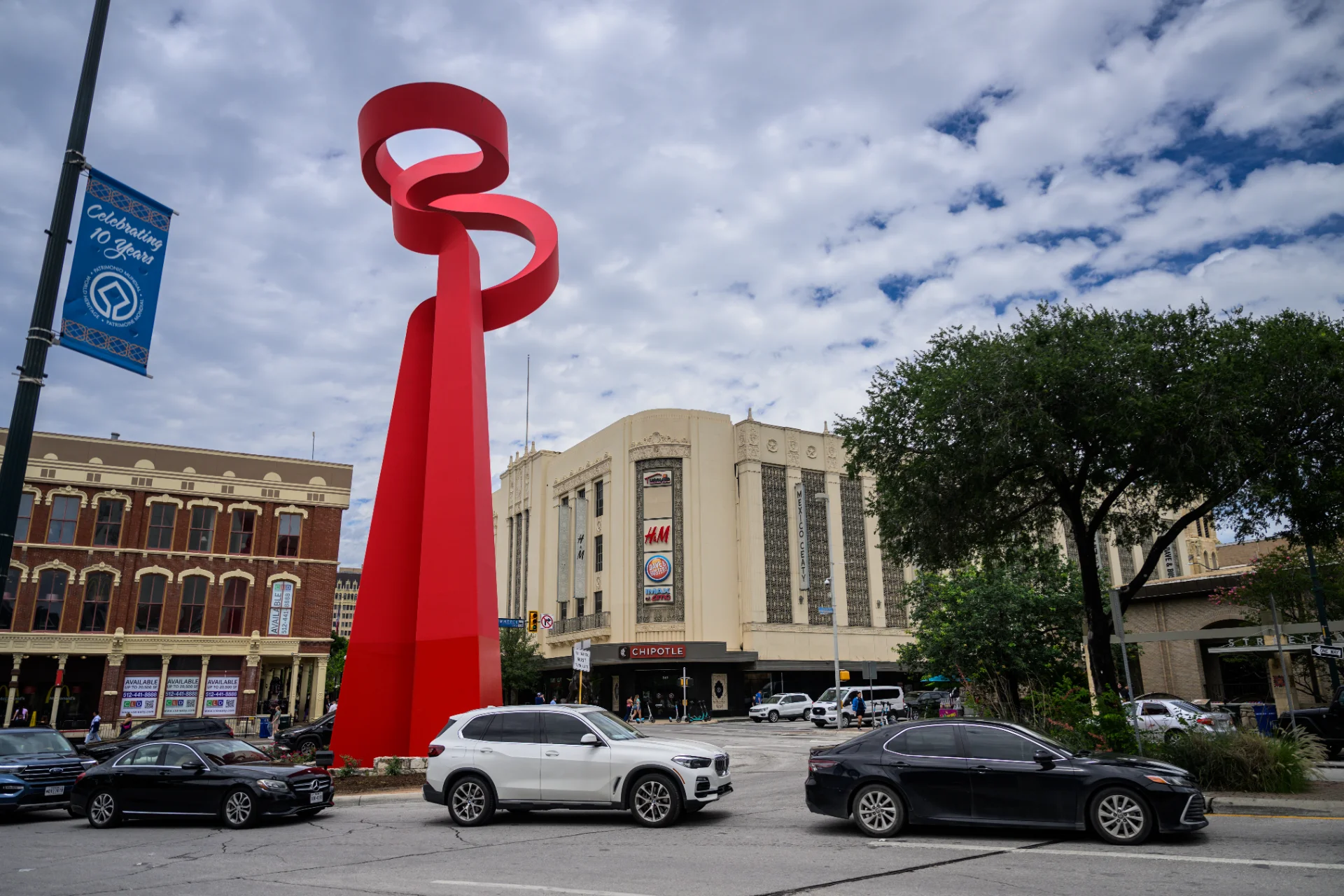 Tall red ribbon sculpture on a downtown San Antonio street corner, with the Shops at Rivercenter building and its H&M, Mexico Ceaty, and Chipotle storefronts in the background