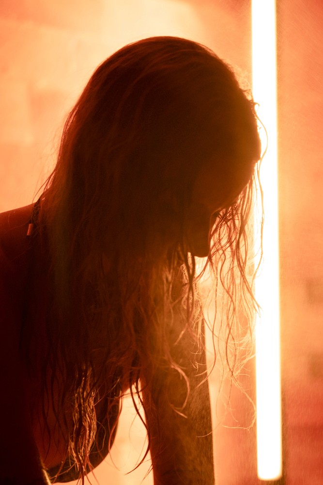 Person facing a warm sauna wall with long flowing hair, bathed in golden light during an aromatherapy sauna session