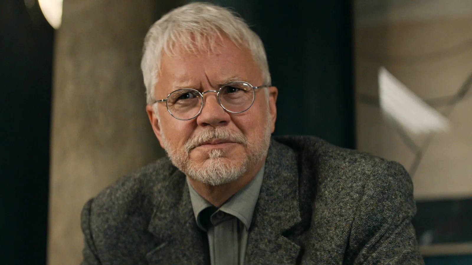 A mature man with grey hair and a beard, wearing round glasses and a grey suit, sits against a blurred background, conveying a serious expression.