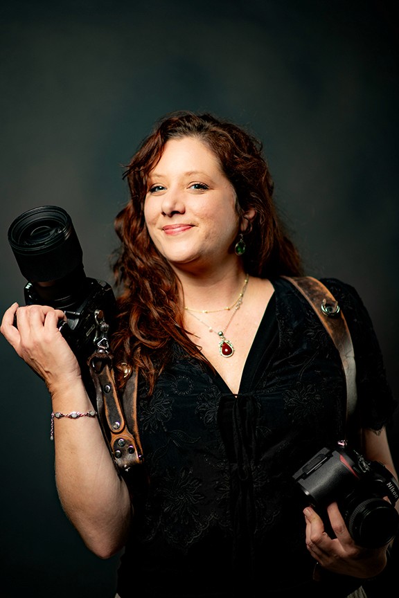Tiffany Teso, photographer, with red hair holding two cameras with leather straps. She wears a black top, and jewelry.