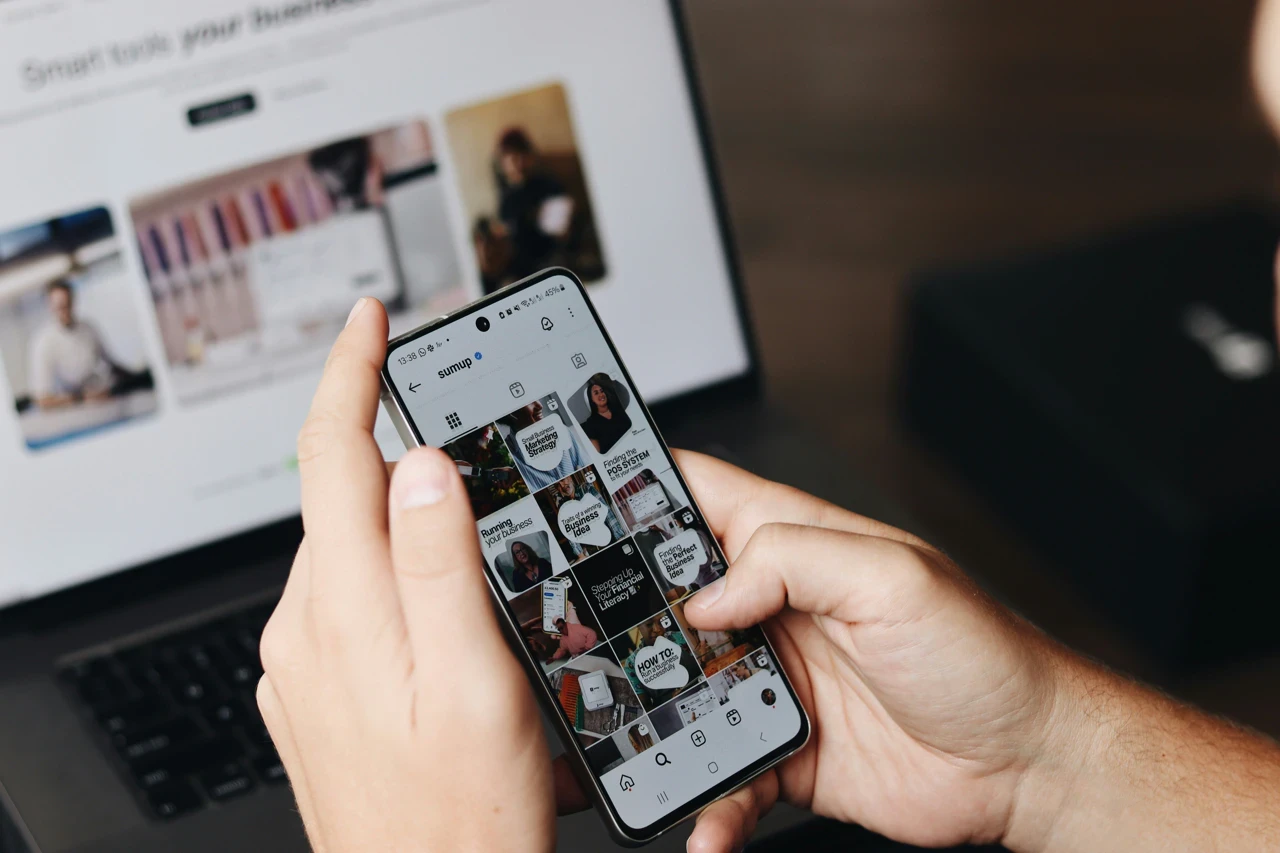 Person hält Smartphone, betrachtet Social-Media-Profil, Laptop im Hintergrund.