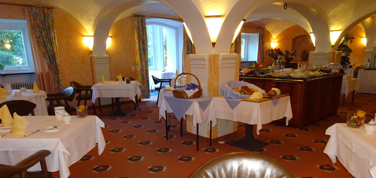 Speisesaal des Hotels: gedeckte Tische mit weissen Tischtüchern unter Gewölbebögen, im Hintergrund ein Frühstücksbuffet.