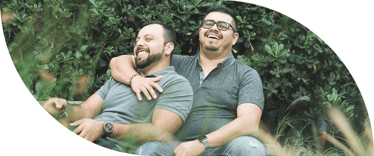 Duas pessoas estão sentadas ao ar livre na grama, sorrindo e rindo juntas, diante de um cenário de folhagem verde exuberante, usando camisetas cinza casuais e aproveitando um momento descontraído.