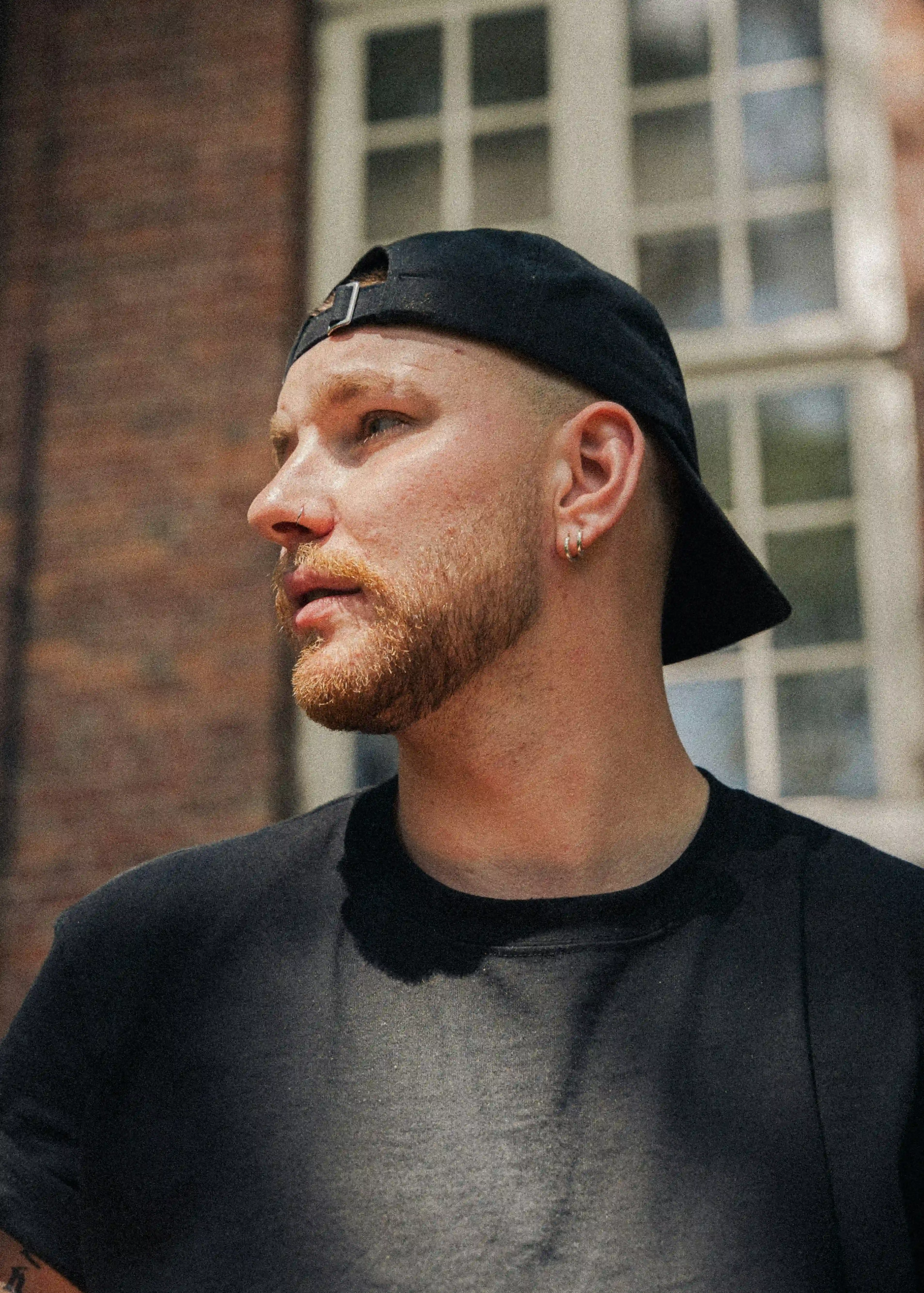 A profile shot of a man with a reddish-blonde beard, wearing a black cap and a black t-shirt, posed in front of a brick building with a window in the background.