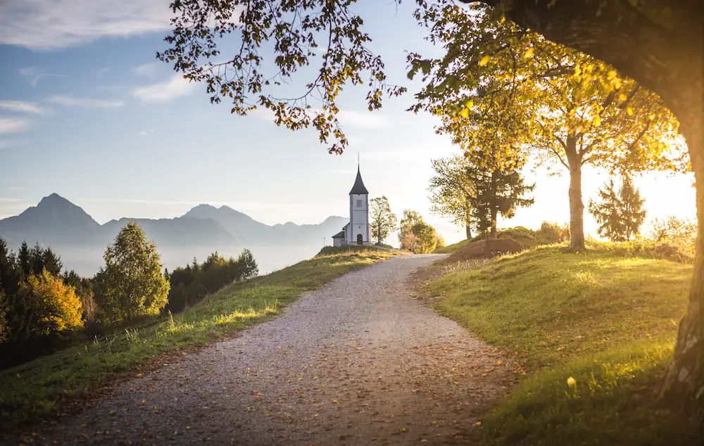The hilltop Jamnik church in Slovenia at sunrise, framed by golden autumn leaves and a view of the distant Alps.