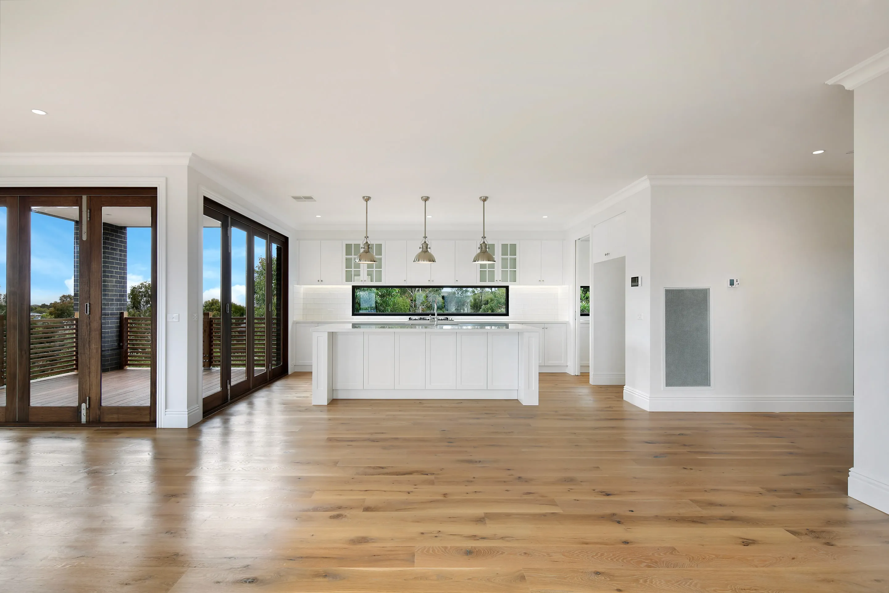 Open living and kitchen with a white shaker island, three nickel dome pendants, a long picture window, oak floors, moulded cornices and timber doors opening to the deck