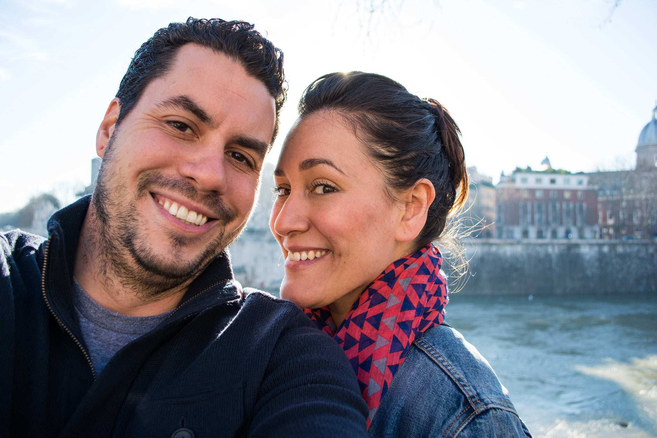 A smiling man and woman in winter clothing pose together outdoors with a river and city buildings in the background.
