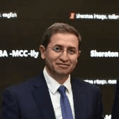 Smiling young woman with curly hair sitting at a laptop, in front of a teal wall.