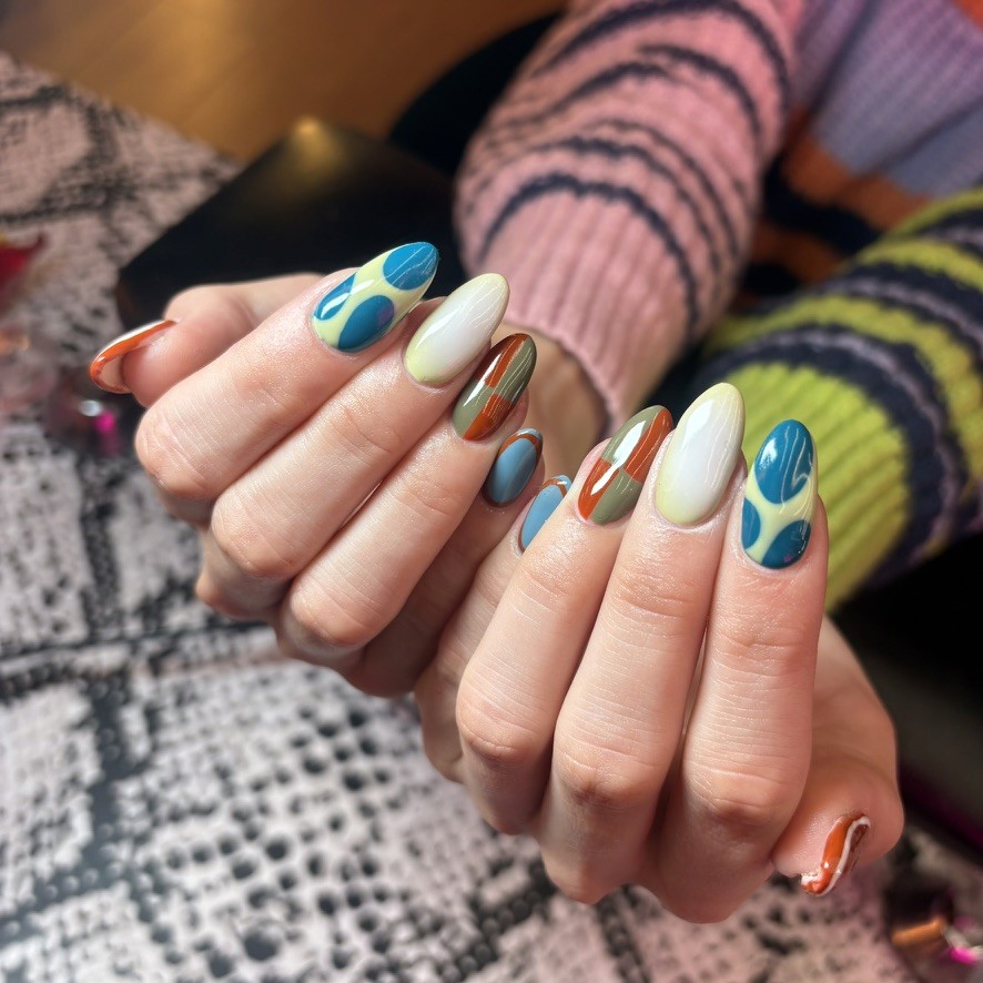 (Full view) Oval nails in a 70s earthy palette of olive, orange, and cream with geometric blocks and teal "lava lamp" teardrop shapes.