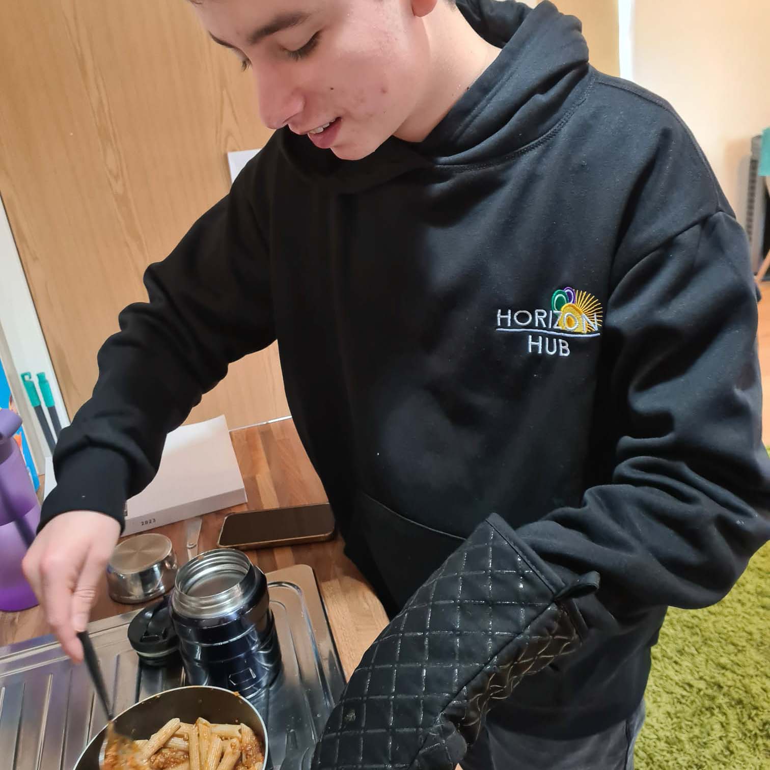 A teenager is cooking using a saucepan, they're wearing an oven mitten and stirring the food