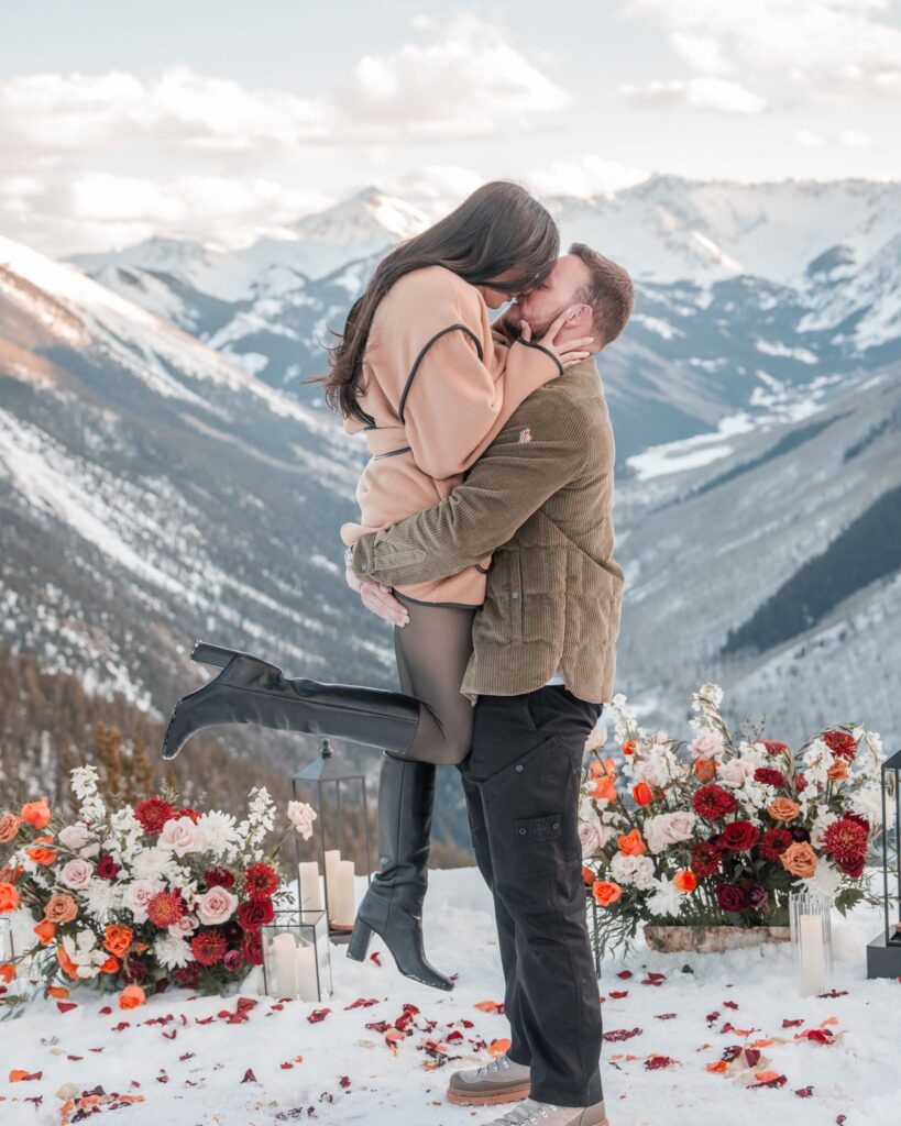 Groom lifting and kissing his newly engaged partner surrounded by floral arrangements and candles at Smith Cabin Aspen, Roaring Fork Valley visible behind them