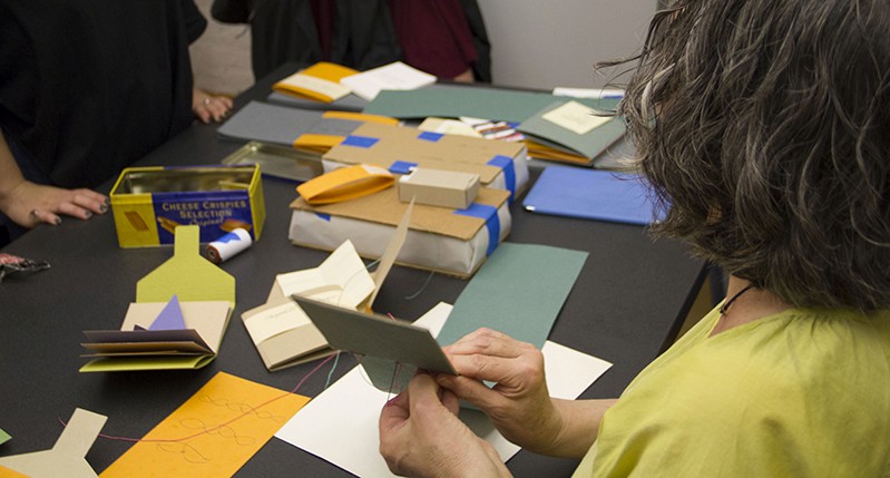 A person working with some colored paper, a needle and a piece of wire, while sitting at a desk.