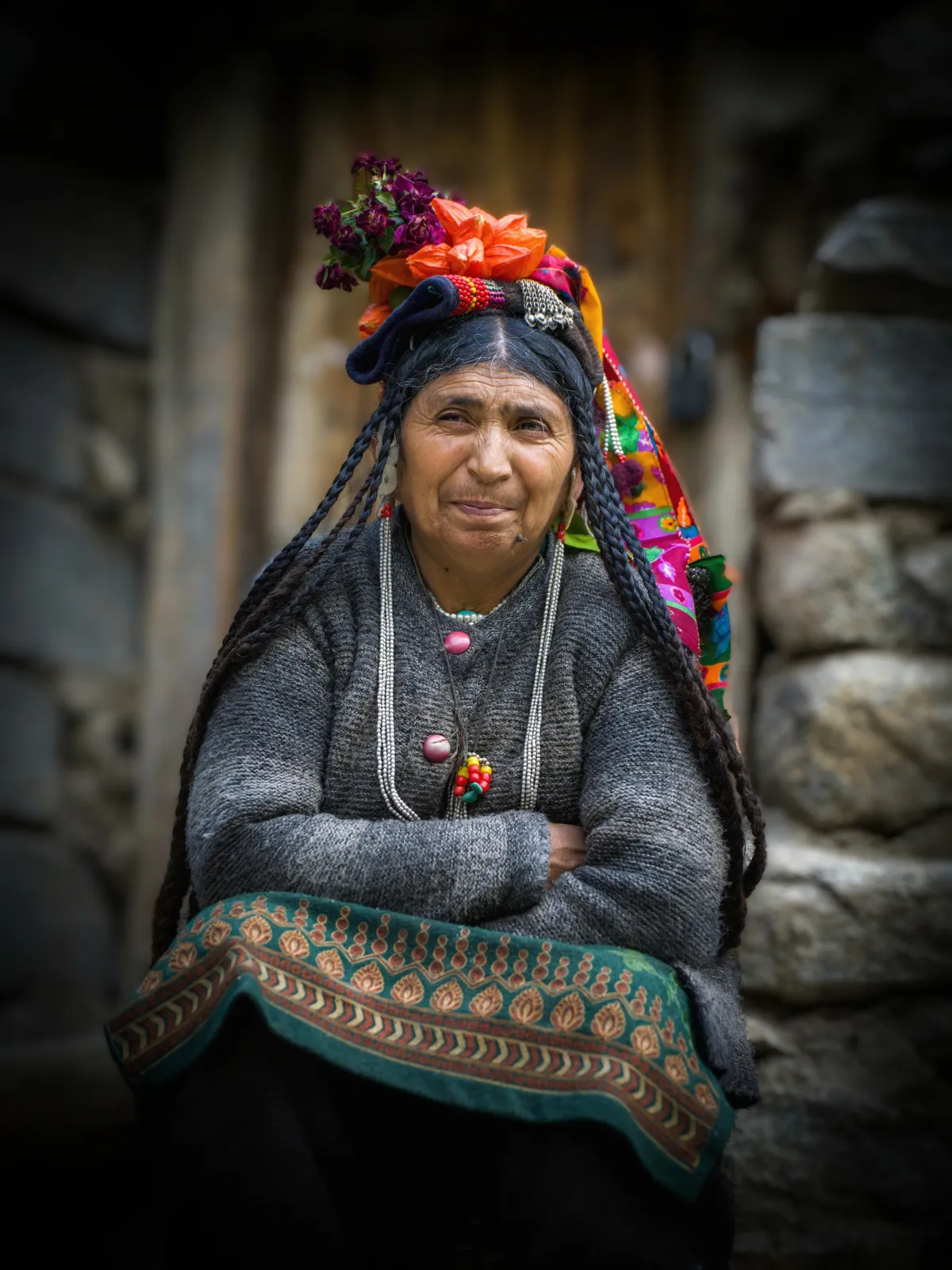 Anciana brokpa con tocado de flores y brazos cruzados junto a un muro de piedra en Dha-Hanu
