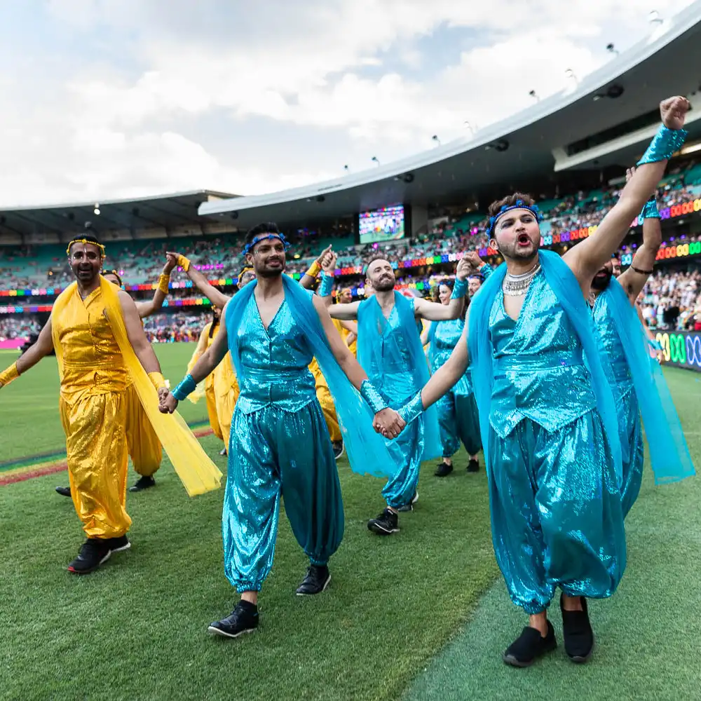 A group of people in shiny blue and yellow costumes dance and hold hands on a stadium pitch, with a colourful audience in the background under a cloudy sky.