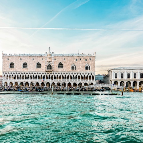 Edificio histórico ornamentado con ventanas arqueadas y columnas junto al paseo marítimo, con barcos atracados delante, bajo un cielo azul despejado.