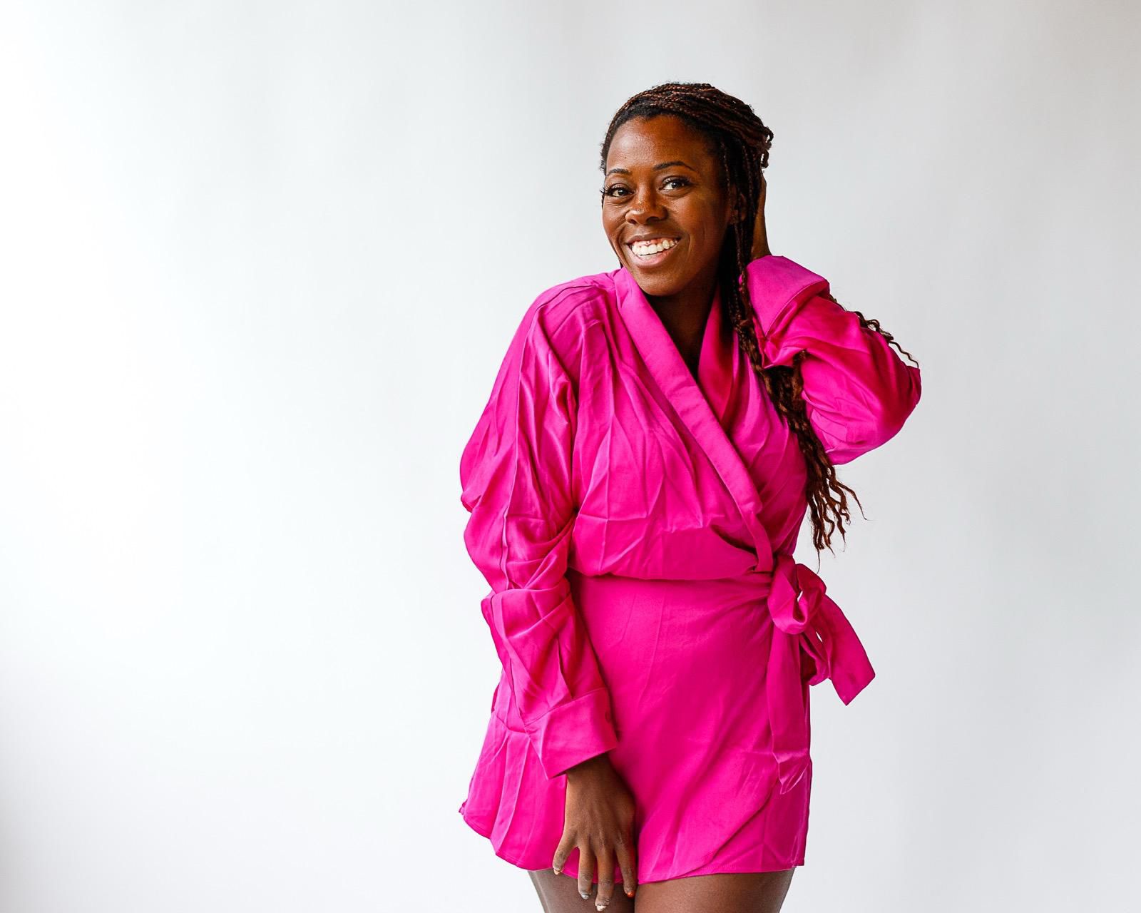 Smiling woman in beige yoga outfit sitting in natural light with green plants in the background.