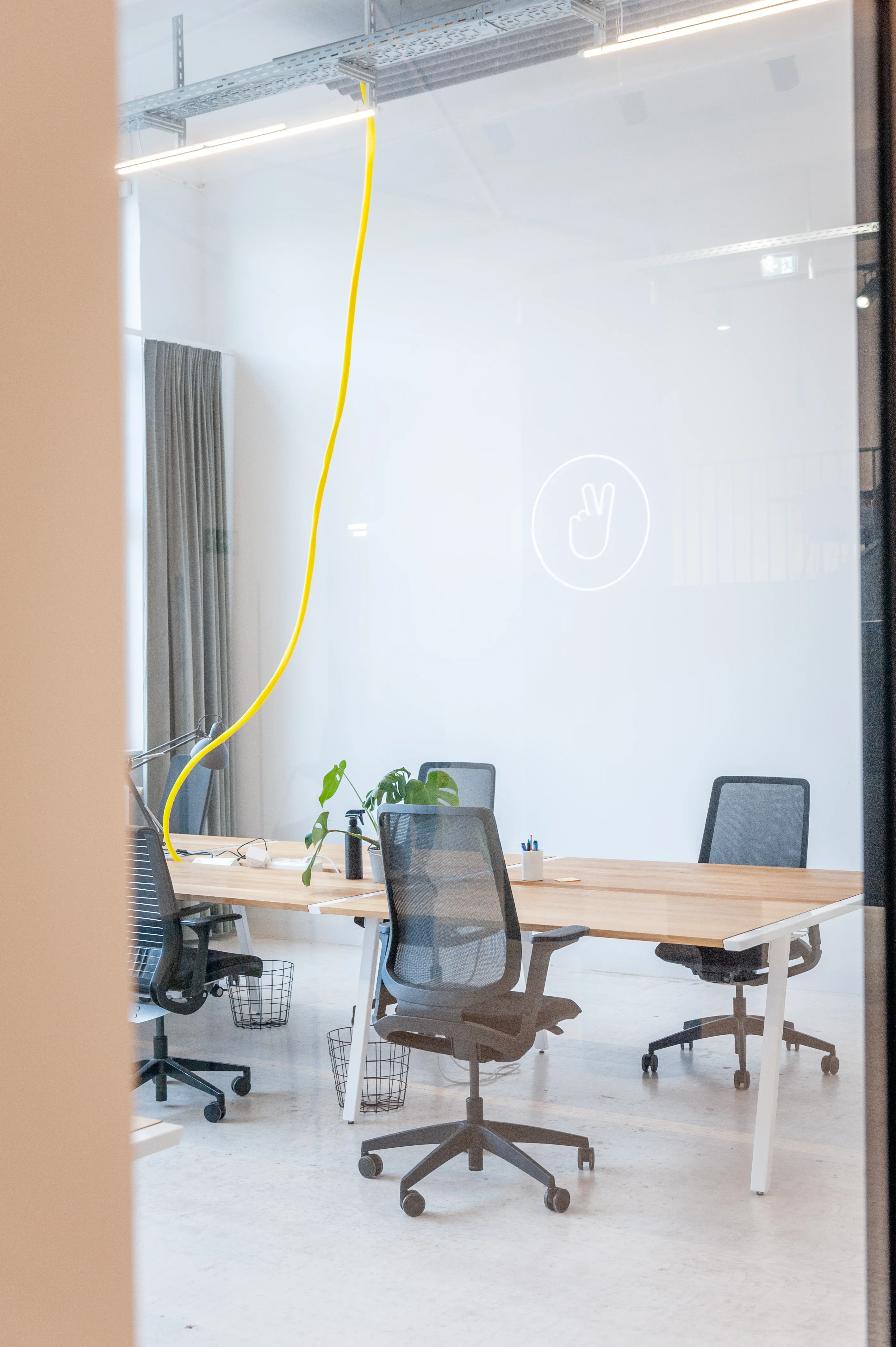 View through a glass partition into a small office at LA RED with white desks, black mesh office chairs, a monstera plant, a yellow cable pendant light and a peace-sign neon on the wall.
