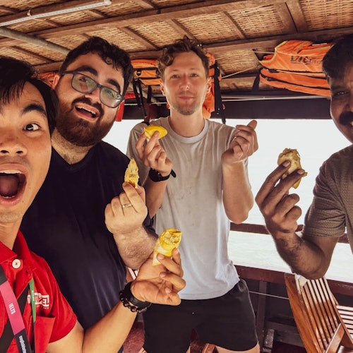 Empat pria dengan gembira memegang potongan makanan di atas perahu kayu, dengan jaket pelampung menggantung di atas mereka.