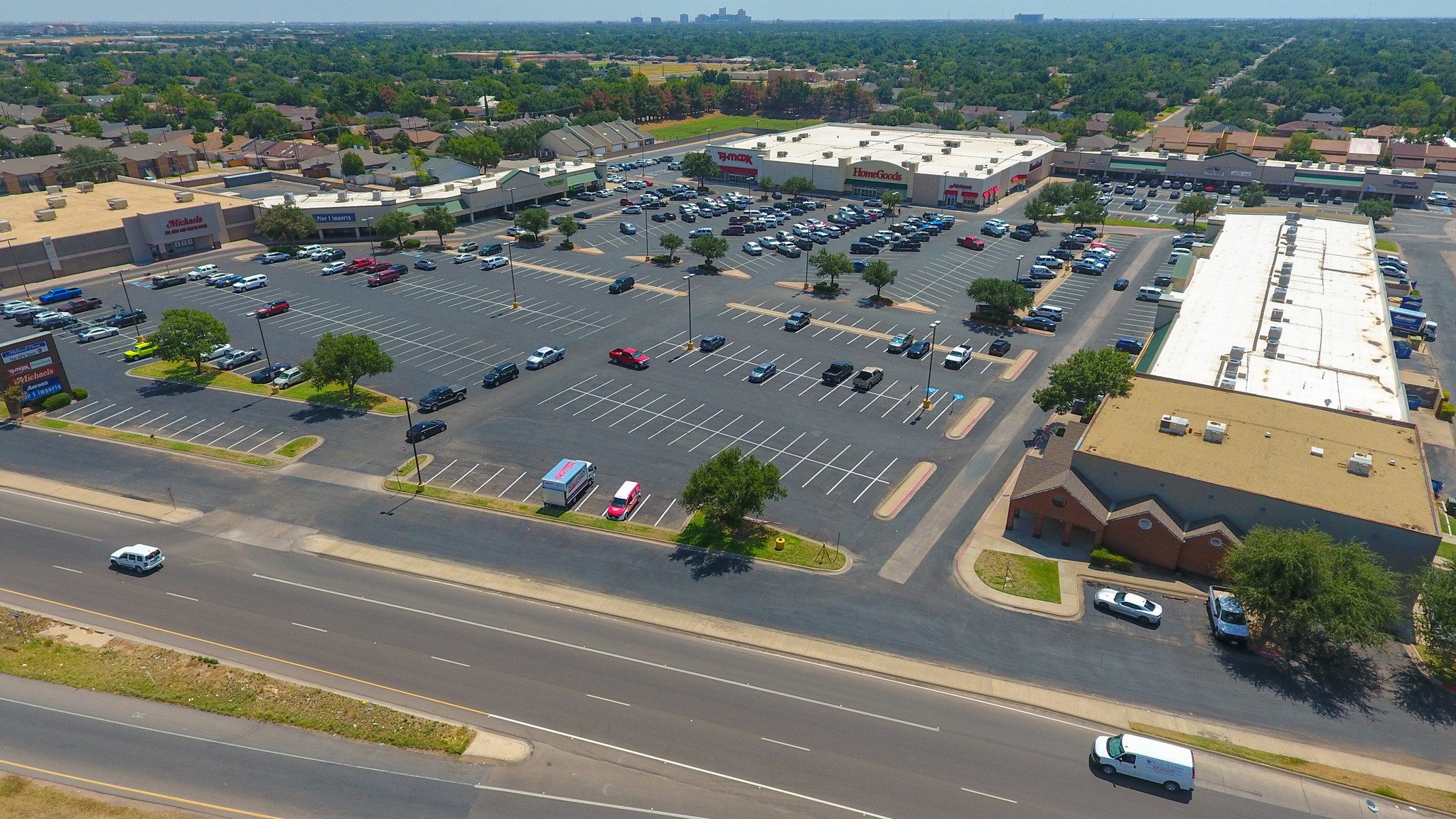 Aerial view of a suburban shopping center with large parking lots, few cars, and scattered trees. Surrounding streets and greenery create a calm, spacious atmosphere.