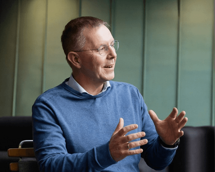 A person in a blue sweater gestures while speaking, seated with a modern interior backdrop.
