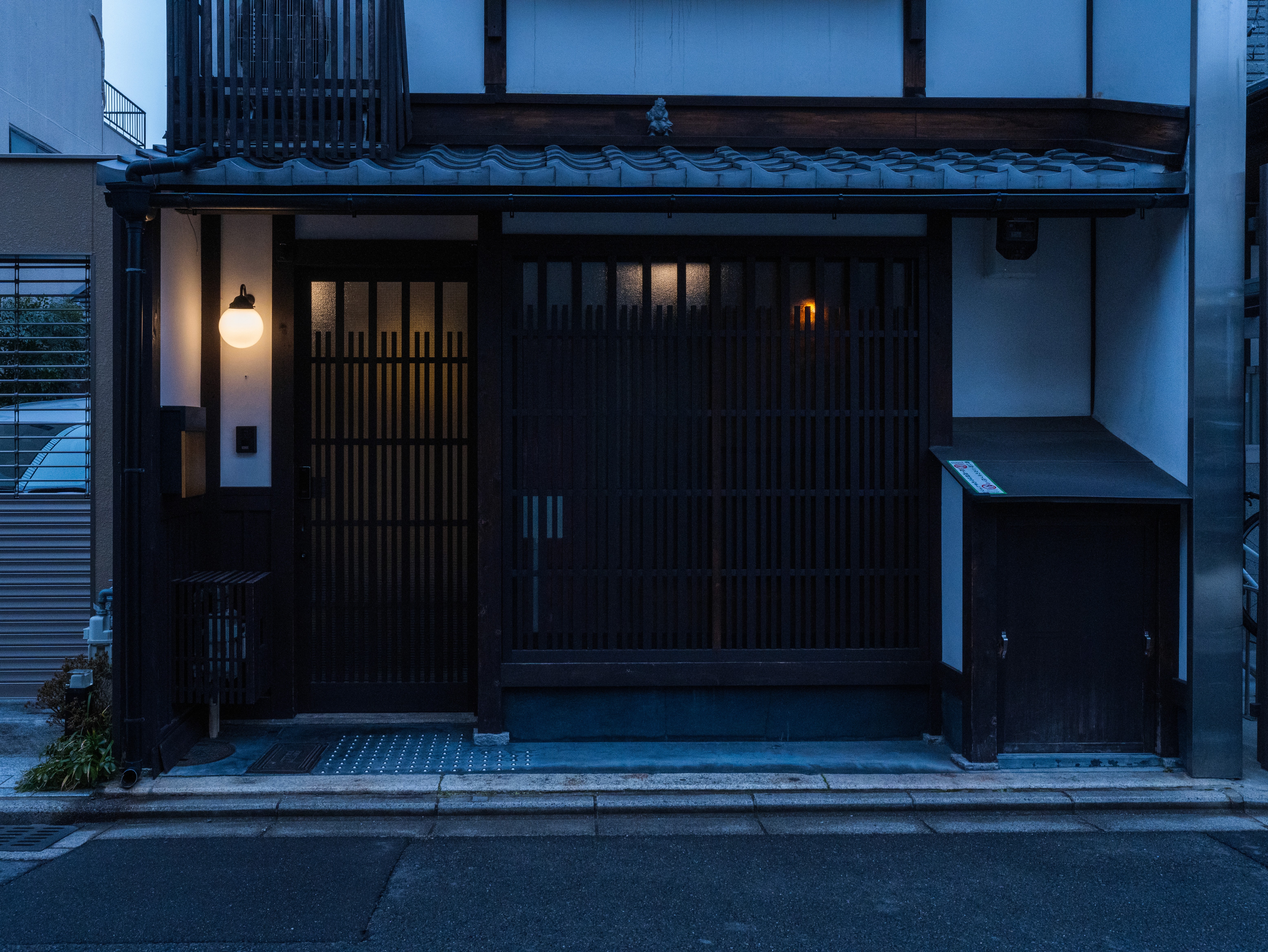 A traditional Japanese building with a dark wood lattice door, illuminated by a single glowing lantern, stands quietly in a dimly lit street.