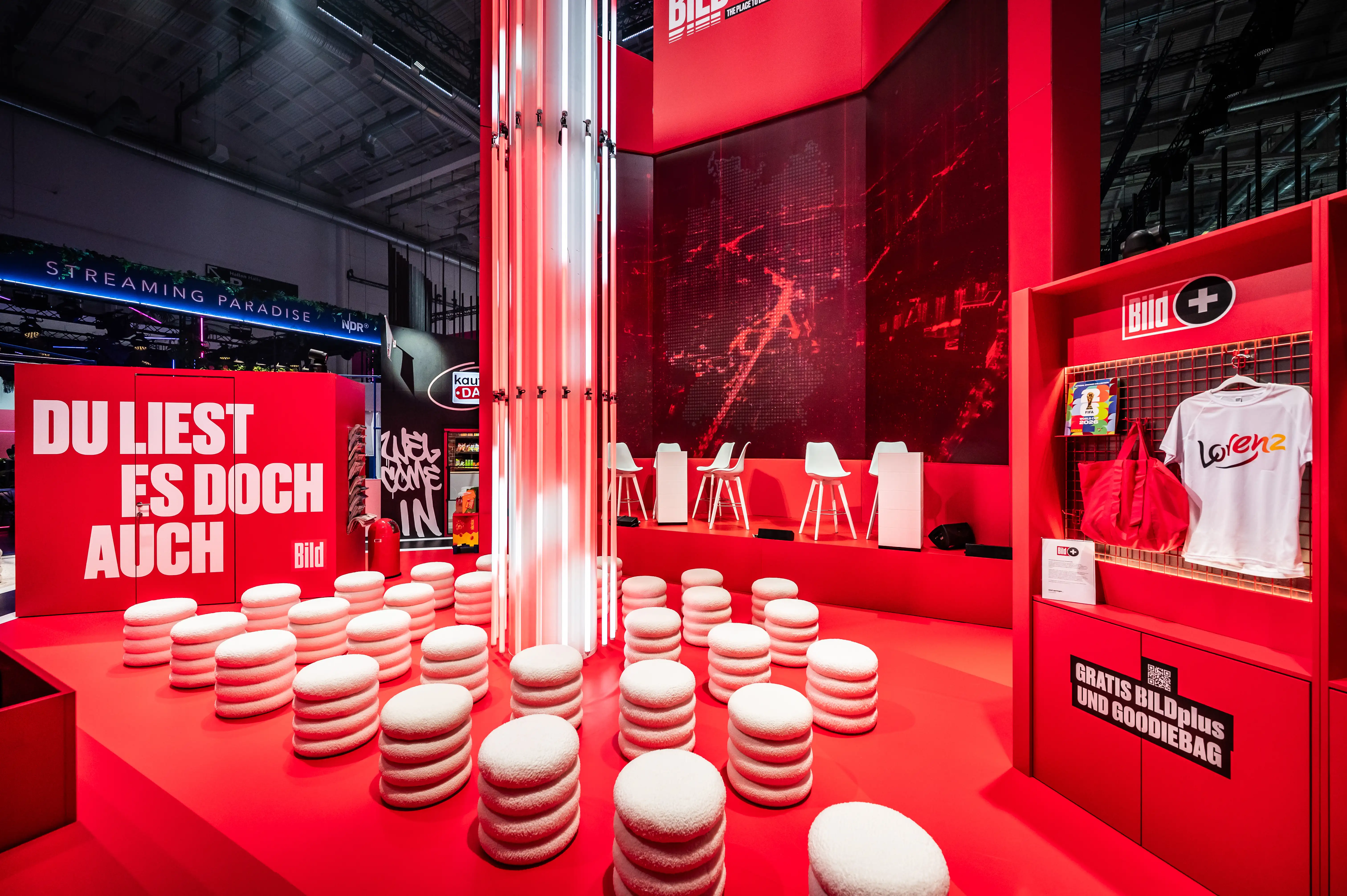 Red Bild lounge with white upholstered stools, slogan wall and merch shelf with shirts and bags.