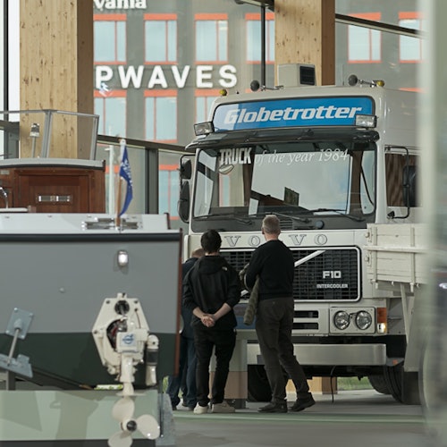 Tre personer står framför en Volvo Globetrotter lastbil inuti en byggnad, med en båt delvis synlig till vänster.