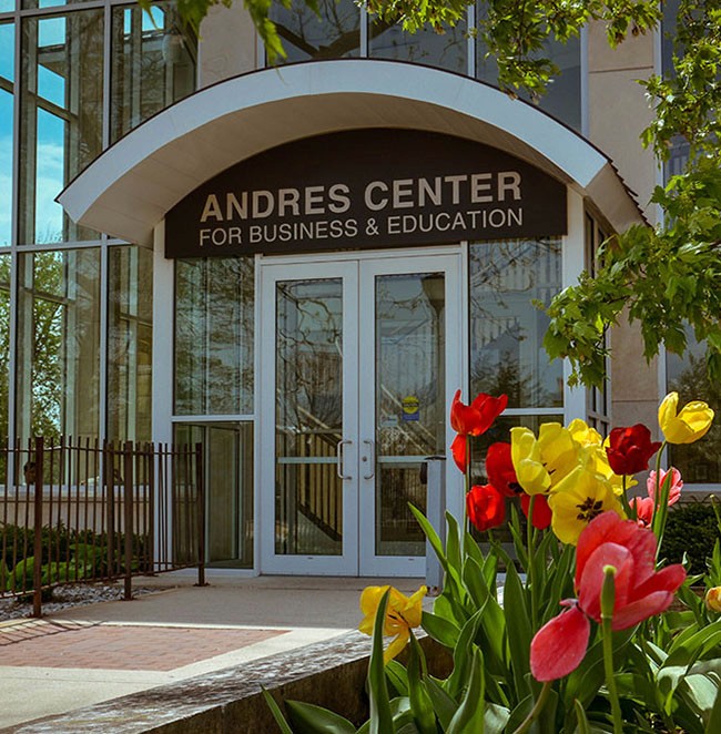 A view of the glassy entrance with colorful flowers in the foreground to a building - the Andres Center for Business and Education