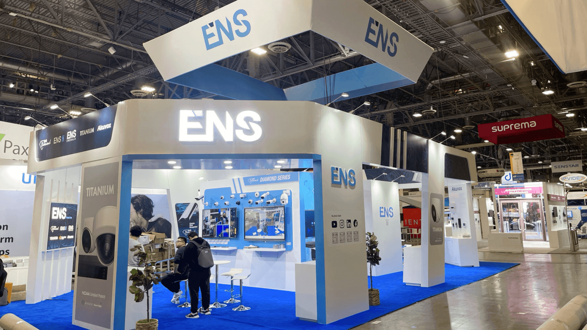 Front product demo area of ENS ISC West 2025 booth with overhead hanging sign, blue flooring, security product displays, and aisle-facing branding