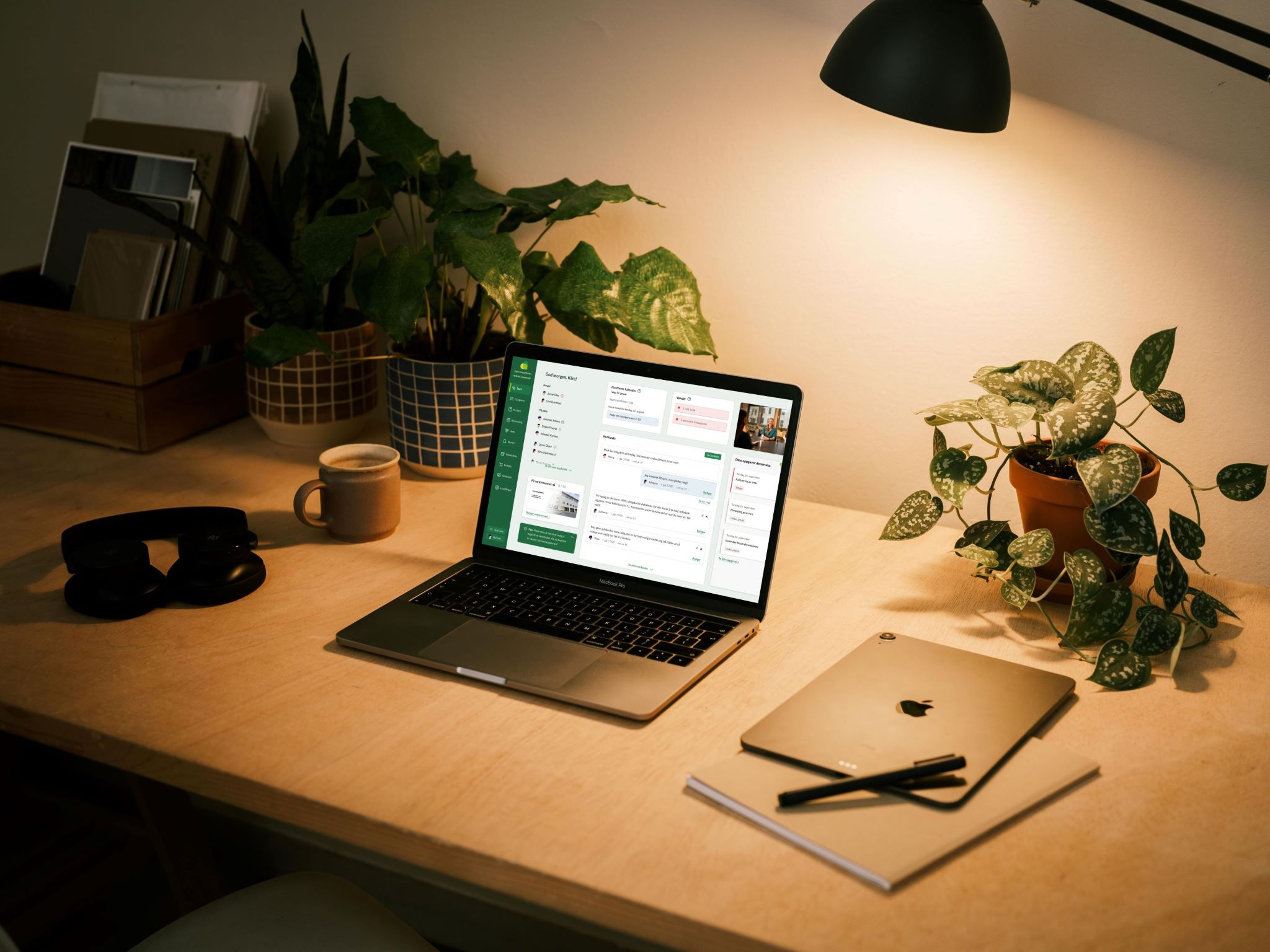 Et koselig arbeidsområde med en bærbar PC, notatbok og planter, opplyst av en skrivebordslampe.
