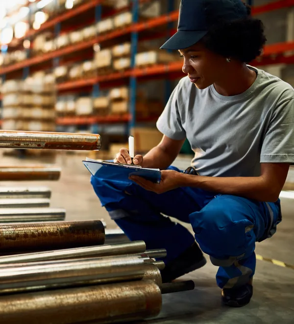 Worker writing on a clipboard while inspecting steel bars stacked in a warehouse