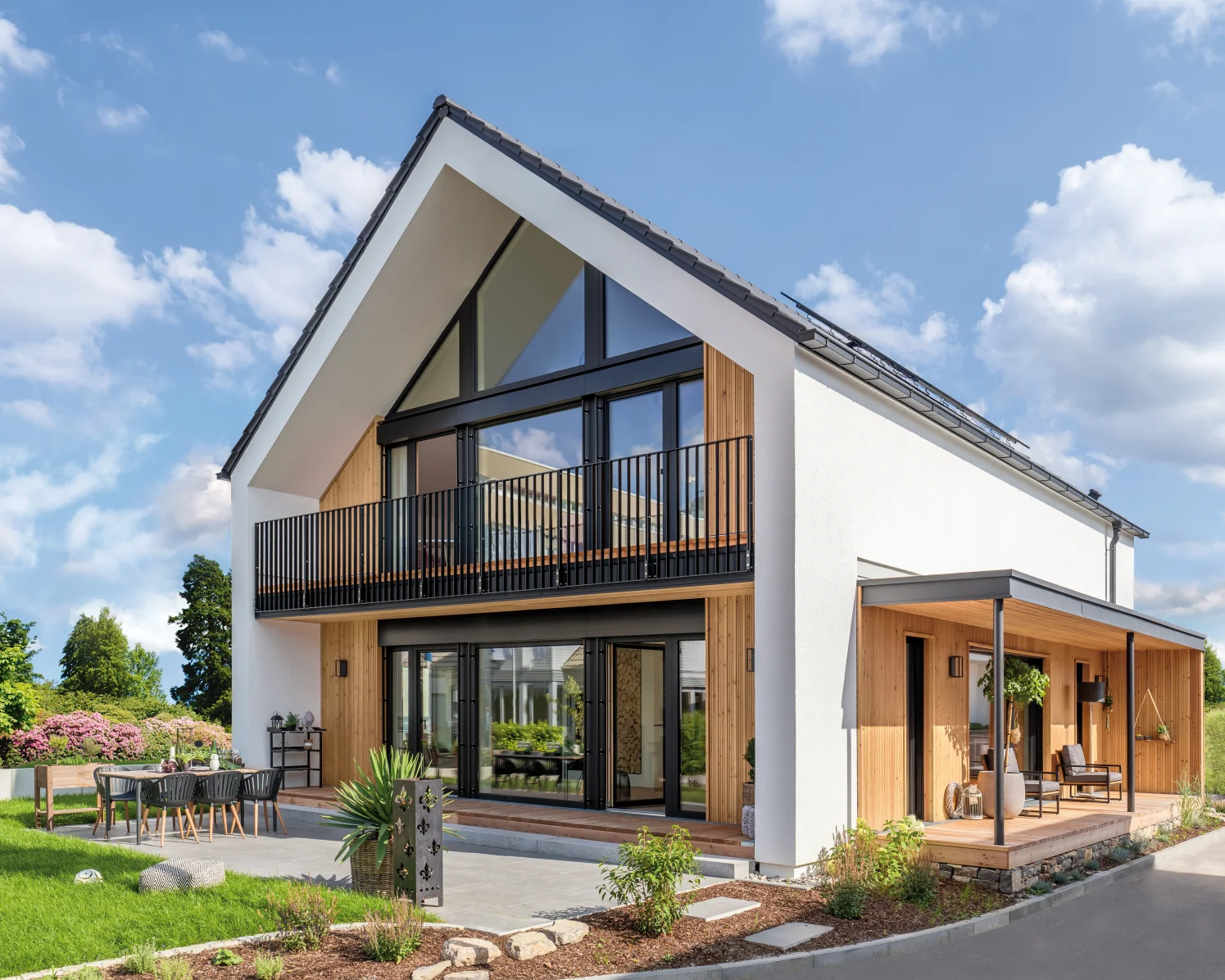 Modernes Einfamilienhaus mit Satteldach, großzügiger Glasfront, Balkon, Terrasse und Gartenmöbeln im Grünen.