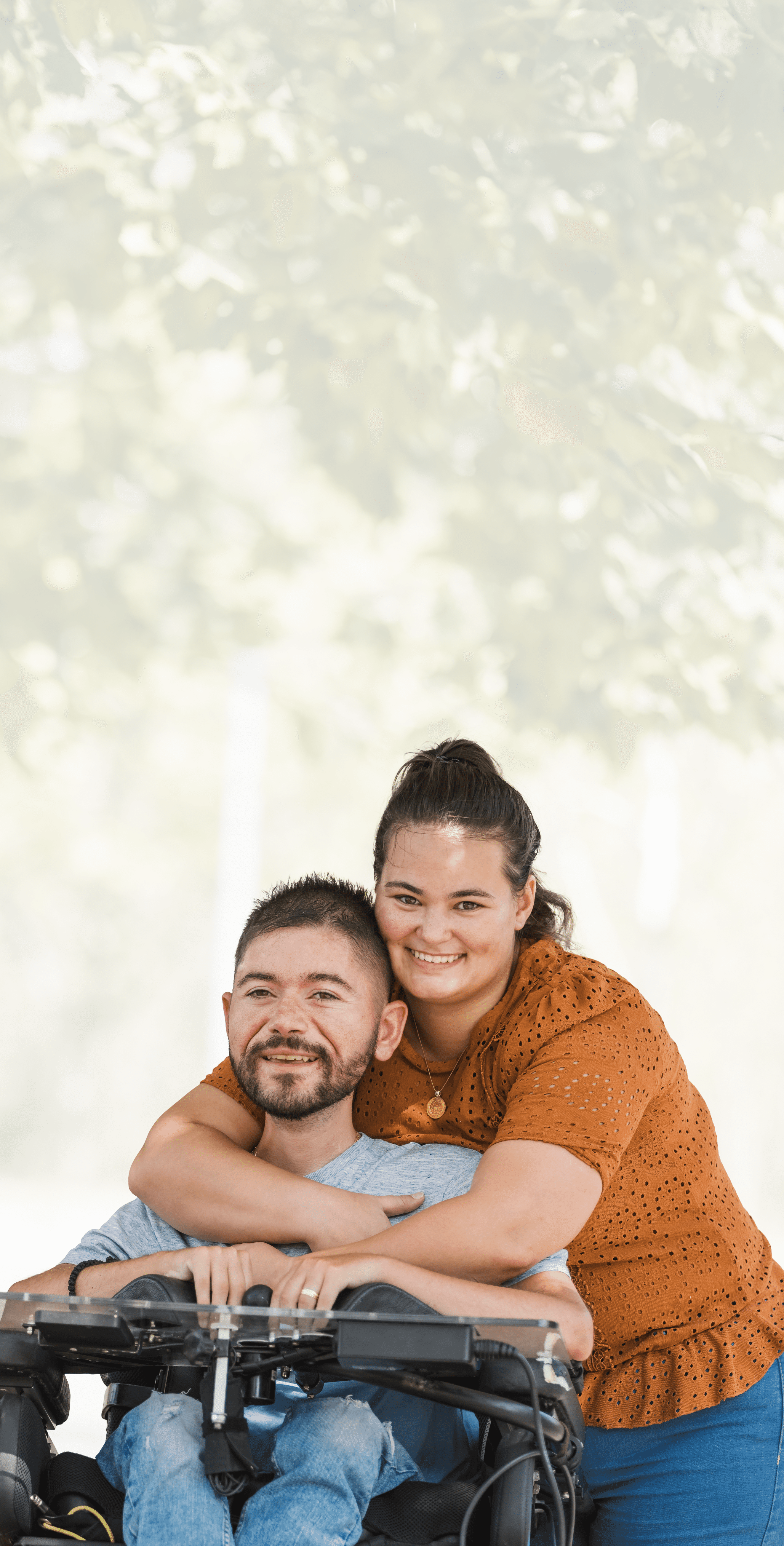 Background Image: A smiling woman hugs a man sitting in a motorized wheelchair outdoors on a sunny day, with green trees and a path in the background. Both appear happy and comfortable together.